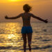 Back view of young happy woman in blue swimwear running with stretched arms into the sea towards colorful sunrise or sunset sky, basking in warm sunlight, rising water splashes