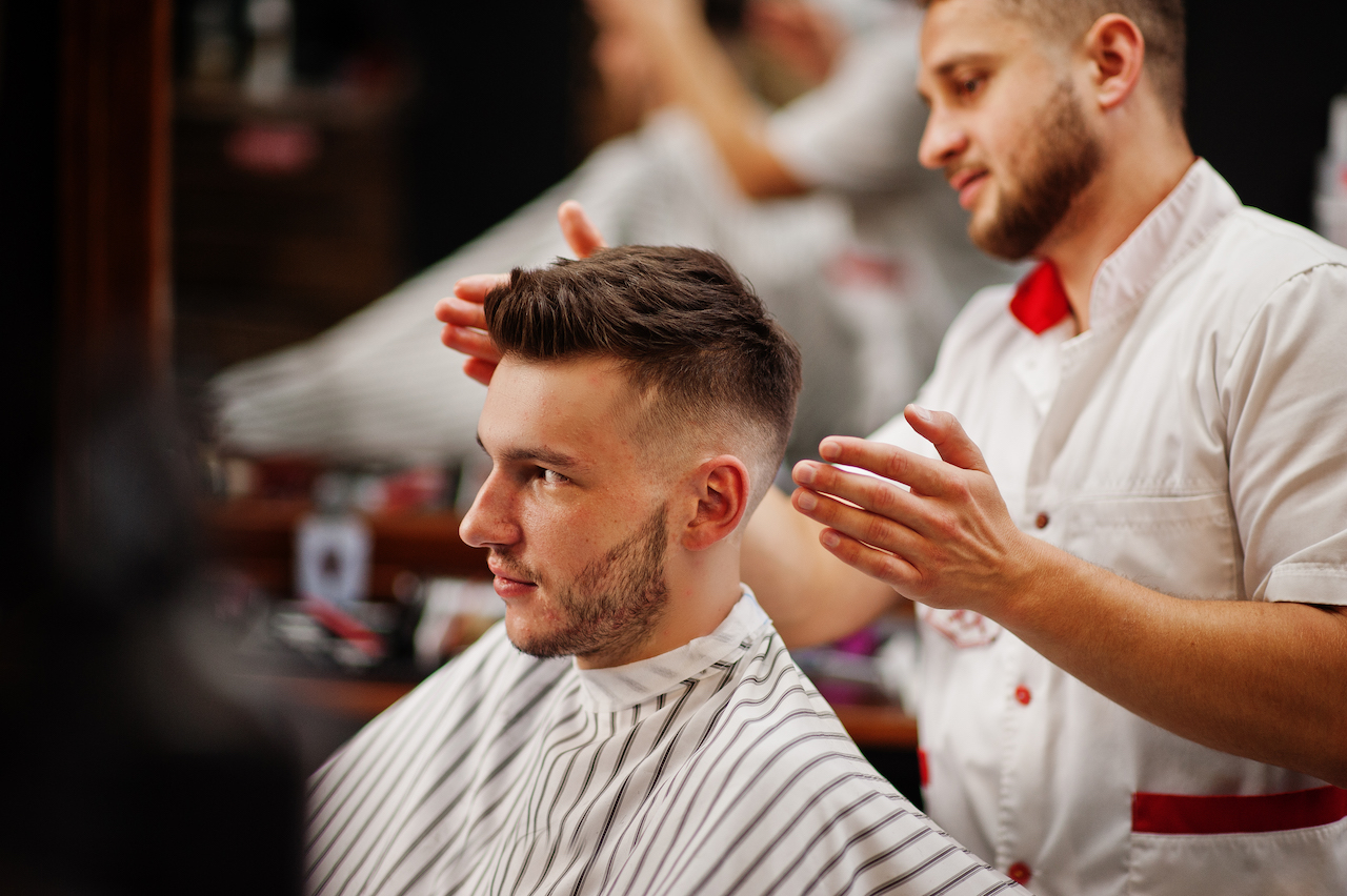 Hombre joven con barba siendo recortado por un barbero mientras está sentado en una barbería.