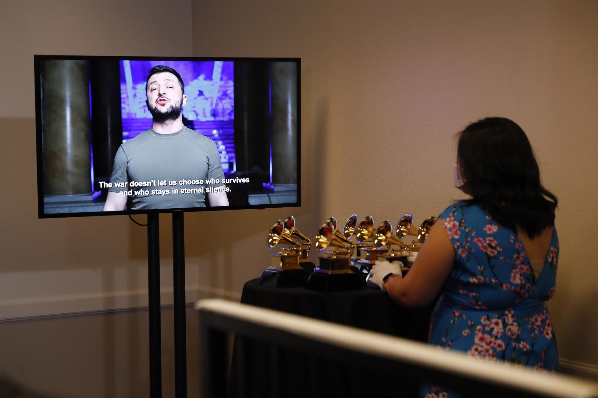 Mujer staff de los Premios Grammy reposa sus manos en mesa con galardones mientras observa el audiovisual de intervención de Volodymyr Zelenskyy en una pantalla. 